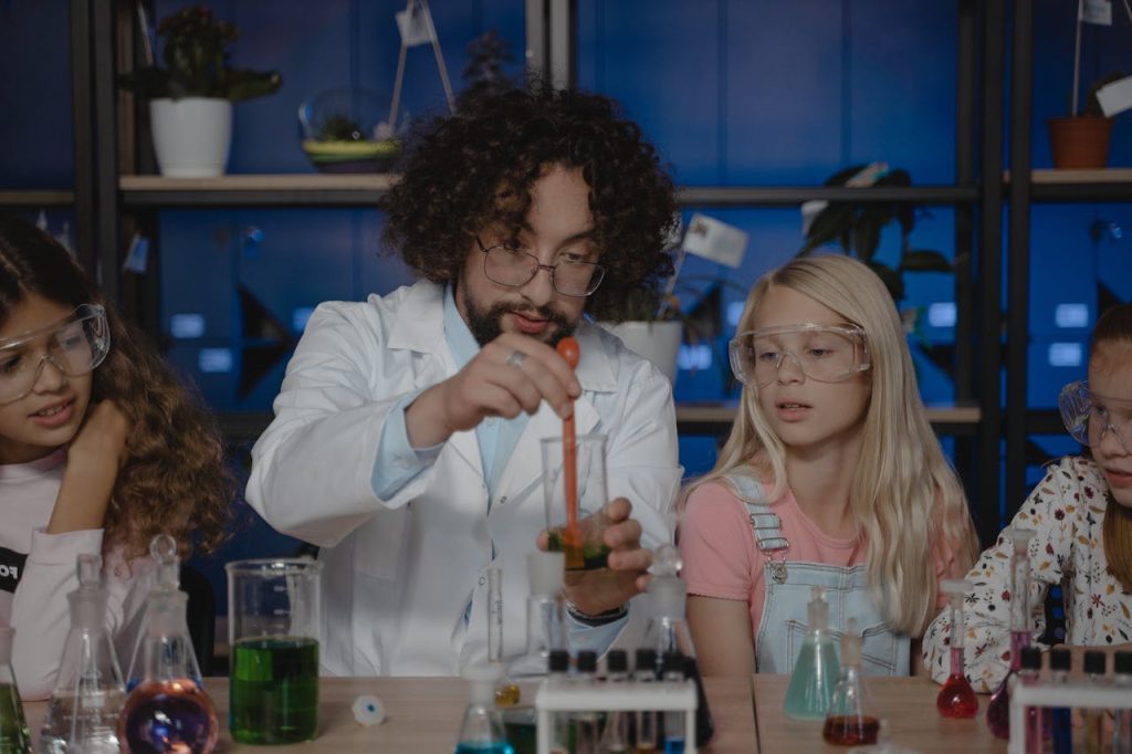 A science teacher in a lab coat demonstrates a chemistry experiment with a beaker and pipette to a group of attentive children wearing safety goggles, surrounded by lab glassware and a periodic table in the background.